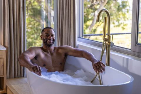 Mature man smiling relaxing in bubble bath near sunlit window with brass faucet