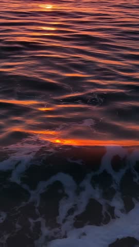Vertical video capturing shimmering ocean waves reflecting descending sun during sunset