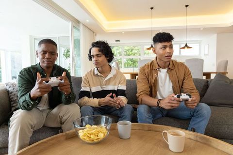 Three friends playing console games on sofa with controllers and snacks modern living room