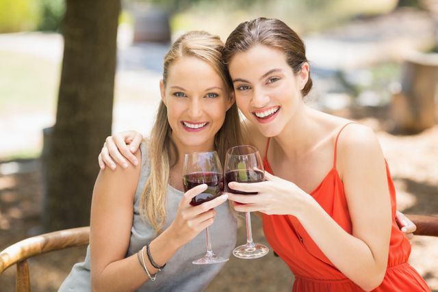 Two women enjoying a sunny day outdoors, toasting with wine glasses. Perfect for use in lifestyle blogs, social media posts about friendship and celebrations, advertisements for wine brands, or promotional materials for outdoor events and gatherings.