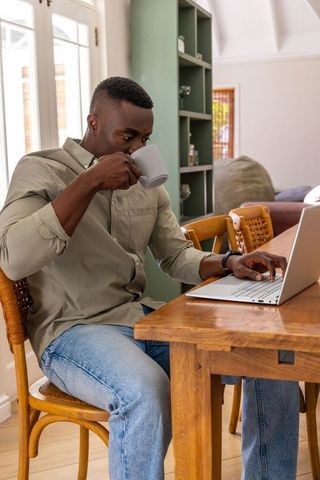 African american man working from home sipping coffee, typing on laptop at dining table