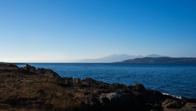 Stunning coastal scenery featuring calm blue waters, rocky shoreline, and distant mountains under a clear blue sky. Perfect for travel blogs, nature websites, relaxation concepts, and environmental campaigns.