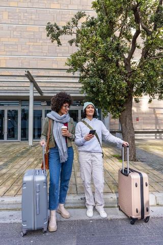 Female friends waiting with luggage outside modern building, chatting and smiling