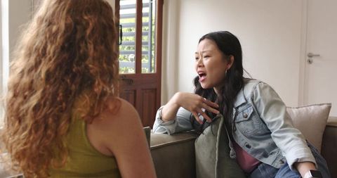 Diverse women chatting on sofa in sunlit living room with denim jacket and smartwatch