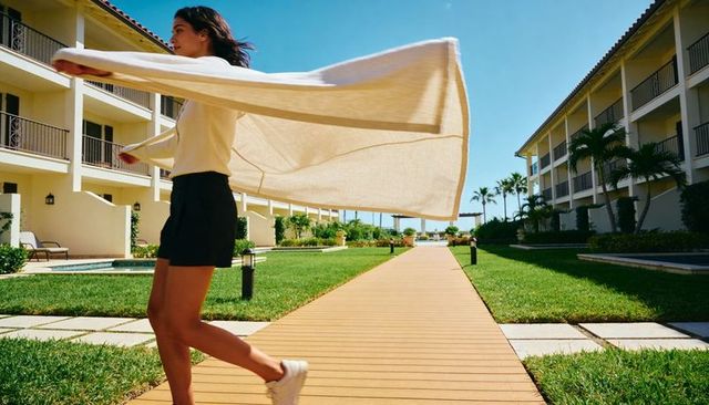 Woman walking resort boardwalk holding billowing beige towel on sunny tropical getaway