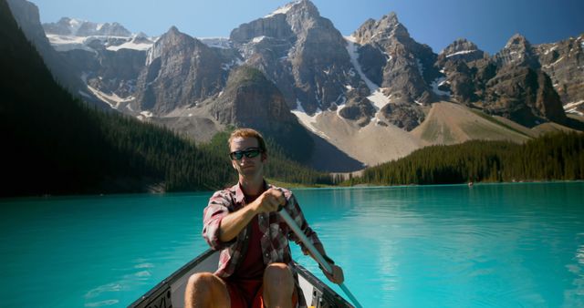 A man is canoeing on a vibrant turquoise lake surrounded by majestic mountains. The clear, tranquil water and stunning scenery create a perfect setting for adventure and relaxation. The sun is shining and there's a peaceful reflection on the lake. Ideal for promotions related to travel, nature tours, adventure activities, and outdoor exploration campaigns.