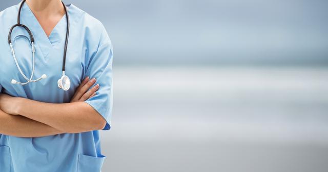 Healthcare professional wearing light blue scrubs standing with arms crossed, with stethoscope hanging from neck, on blurred background. Ideal for themes on medical services, nurse appreciation, hospital environment, healthcare promotions, patient care, and professional healthcare workforce.