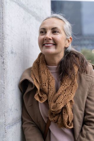 Senior woman smiling, leaning on concrete wall in urban plaza wearing brown coat and knit scarf