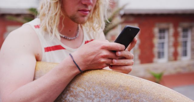 Young man with blonde hair holding surfboard in urban area, checking smartphone. Ideal for themes about youth, surfing, technology, and outdoor activities.