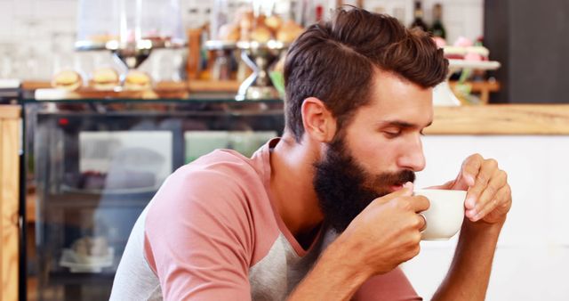 Man with beard drinking coffee in cozy cafe setting. Represents relaxation, casual lifestyle. Perfect for articles on trendy cafes, relaxation tips, fashion blogs, or lifestyle content.