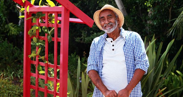 Elderly man enjoying time in garden, wearing a straw hat and casual outfit, standing near red garden structure with green plants. Ideal for themes involving gardening, outdoor activities, senior lifestyle, leisure time, and nature. Can be used in advertisements, blogs, or articles related to healthy aging, hobbies, and wellness.