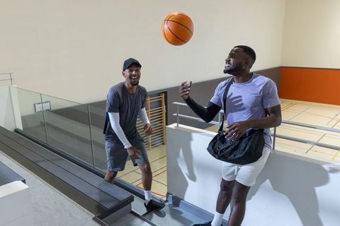 Two men tossing basketball and laughing on gym balcony carrying duffel bag