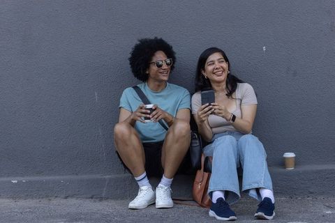 Two friends laughing on urban ledge holding coffee and smartphones, casual street lifestyle