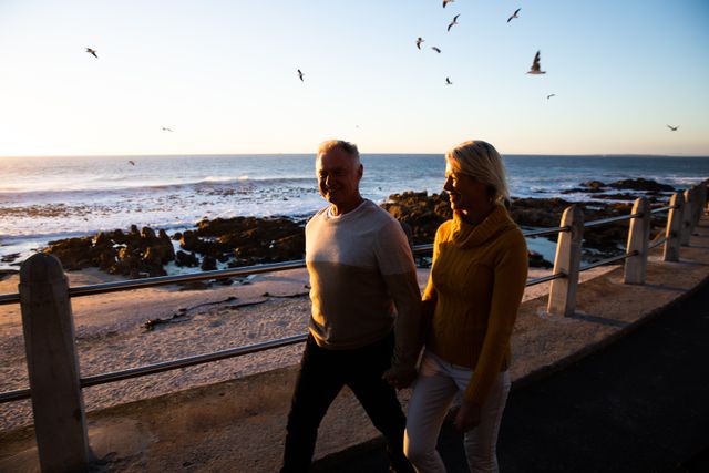 Front view of a happy senior Caucasian couple in love enjoying time in nature together, walking on a promenade, holding hands, during a sunset