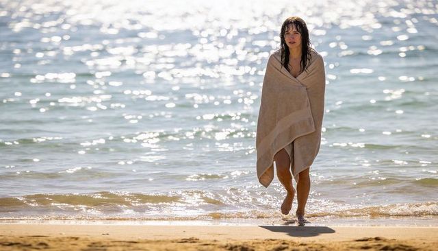 Woman wrapped in towel walking from sparkling sea onto sunlit sandy shore, barefoot and calm