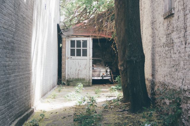 Classic car parked in a rundown garage, with overgrown vegetation and natural light filtering in. Scene depicts urban decay and neglect, with elements of vintage and retro. Perfect for concepts such as forgotten history, nature reclaiming urban spaces, or automotive enthusiasts.