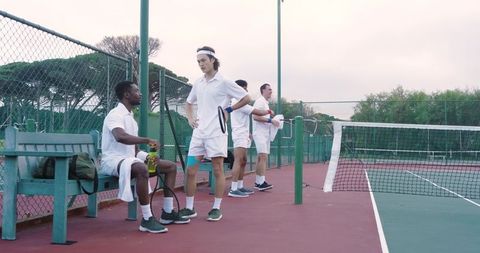 Diverse male tennis players resting courtside on outdoor hardcourt during club practice