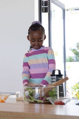 African american girl preparing fresh salad in bright kitchen wearing pastel striped top