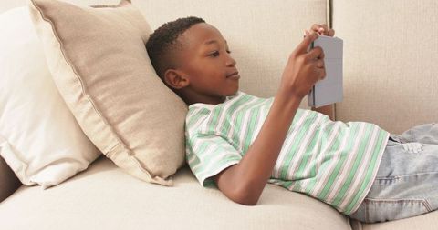 Boy reclining on sofa using tablet for gaming and learning, relaxed home technology scene