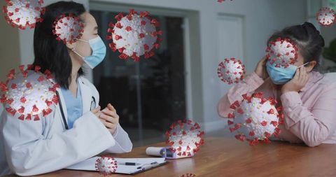Doctor consulting patient wearing masks at desk with floating coronavirus illustration