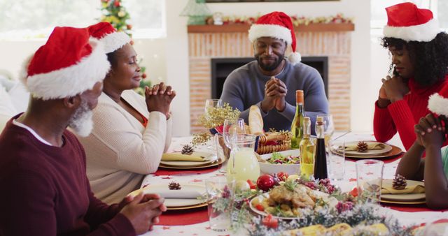 Family wearing Santa hats, holding hands and praying before a holiday meal. Christmas decorations and food on table. Ideal for holiday season promotions, family gatherings, and festive advertisements.