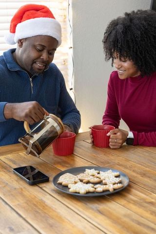 African american couple enjoying holiday coffee, man in santa hat pouring french press