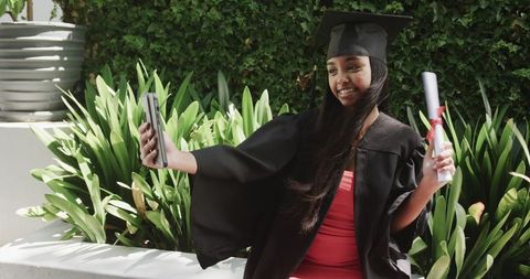 Young adult woman in graduation gown taking selfie with diploma in garden courtyard