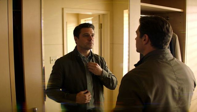 Man adjusting collar while checking mirror in compact dressing area wearing olive jacket