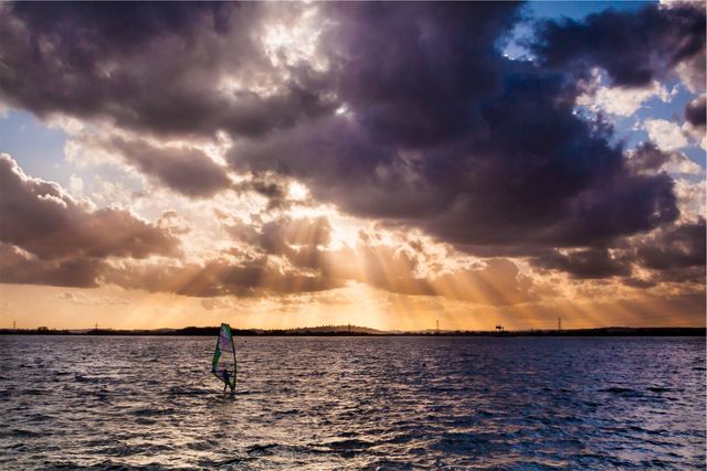 This image showcases a windsurfer navigating the ocean during sunset, with dramatic clouds and rays of sun breaking through. Ideal for promoting adventure sports, travel destinations, and active lifestyles. Perfect for use in advertising, blogs, website headers, and magazines focused on water activities or nature photography.