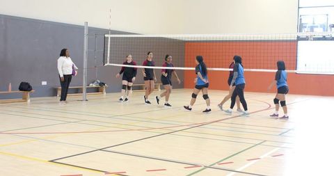 Diverse female volleyball teams gathering near net in indoor gym with coach holding ball