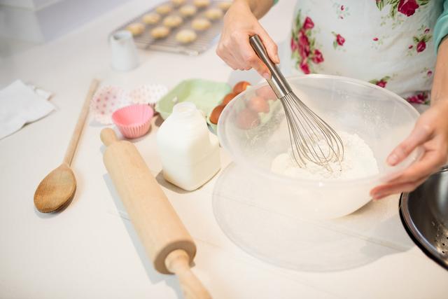 Woman whisking flour in a bowl in a kitchen, surrounded by baking ingredients and tools. Ideal for content related to home baking, cooking tutorials, recipe blogs, and kitchenware advertisements.