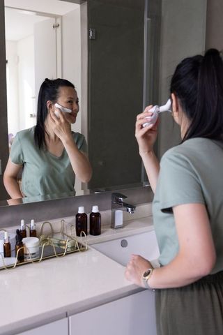 Woman using handheld facial brush at modern bathroom vanity for skincare routine
