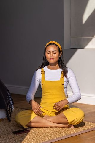 Sunlit mindfulness at home: asian woman meditating in yellow overalls, cross-legged calm