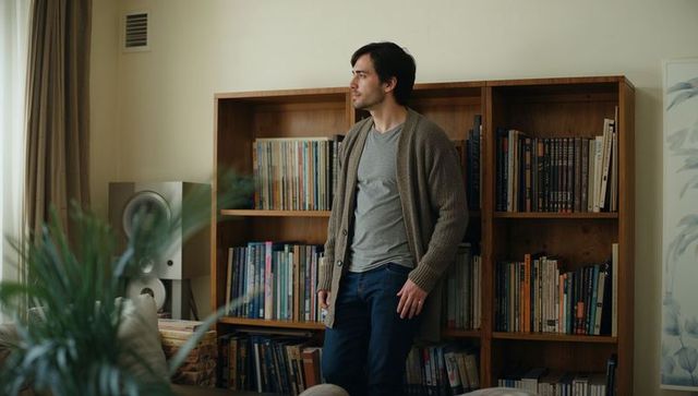 Contemplative man leaning by bookshelf wearing knit cardigan and gray tee in sunlit room