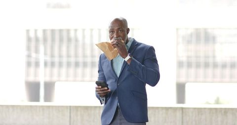 Mature African American Executive Eating Sandwich While Checking Phone on Office Ledge