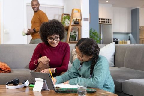 Black mother helping daughter with homework on couch using tablet and notebook in cozy living room