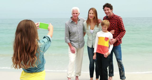 Happy family standing on the beach, with the ocean in background, while young girl taking photo, and capturing memories during vacation. Useful for advertising family holidays, promotions of family bonding, beach vacations, travel marketing, and lifestyle features.