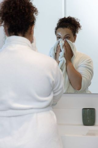African american woman practicing skincare holding towel at bathroom mirror in white bathrobe