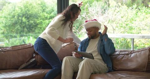 Couple celebrating holiday on sofa sharing tablet and laughing with festive hats