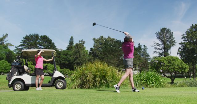 Two women are engaged in a game of golf on a bright sunny day. One woman is preparing to take a shot while the other watches from a golf cart. The scene is vibrant with green trees and a clear sky. This image can be used in promotions for golf courses, sports apparel, outdoor activities, and vacation packages.