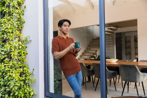 Asian man enjoying coffee by sliding glass door in modern open-plan dining with staircase