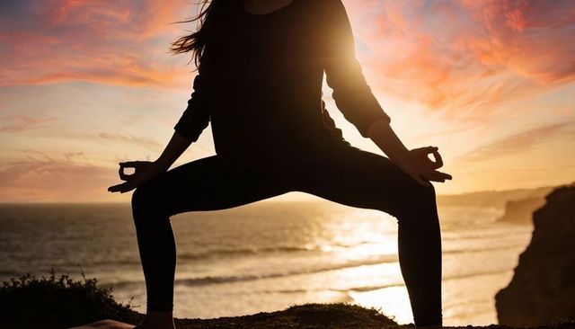 Silhouetted woman practicing wide squat yoga on coastal cliff during vibrant sunset glow