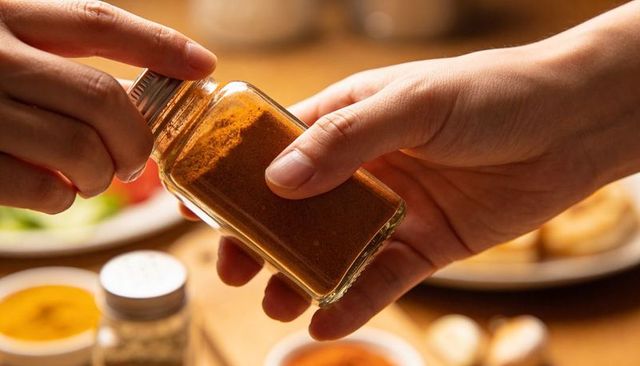 Hands twisting metal lid off glass spice jar over wooden table holding paprika seasoning