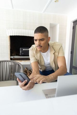 Man concentrating on smartphone while working on laptop in bright sunroom with fireplace