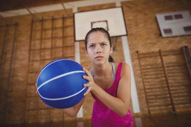 High school girl holding basketball with determined expression in gym. Ideal for content related to youth sports, athletic training, school activities, teamwork, and fitness. Can be used in educational materials, sports promotions, and motivational content.