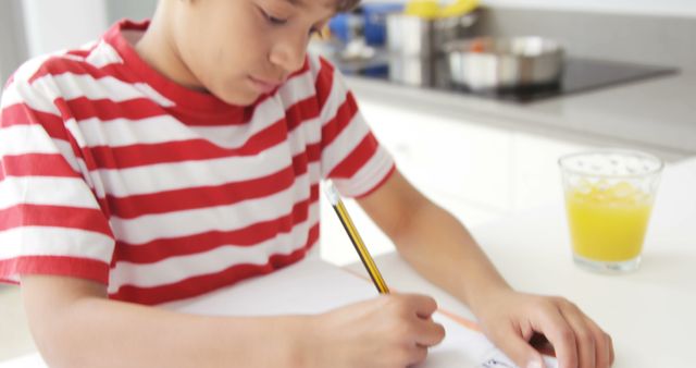 Young boy concentrating on homework at kitchen table, writing in notebook with pencil. Glass of orange juice on table. Suitable for educational materials, family lifestyle themes, and child development concepts.