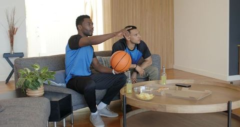 Multicultural friends watching basketball game at home holding ball and pointing