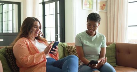 Friends sharing moment on sofa, smiling and using smartphones in bright modern living room