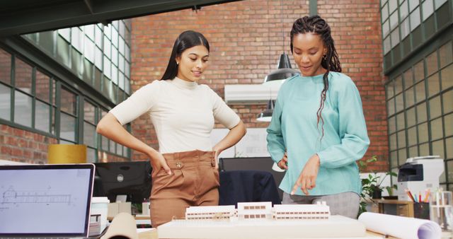 Two female architects in a modern office discuss an architectural model placed on a desk. They stand close together, considering design plans and discussing project details. The background consists of a brick wall and large windows, imparting an industrial chic look. This image can be used in articles or advertisements related to architecture, female professionals, teamwork, and creative workspaces.