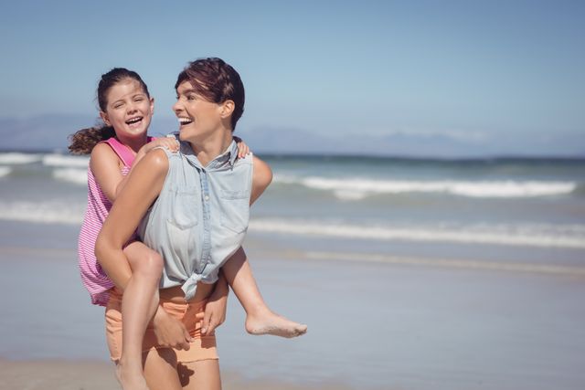 Cheerful mother piggybacking daughter at beach during sunny day